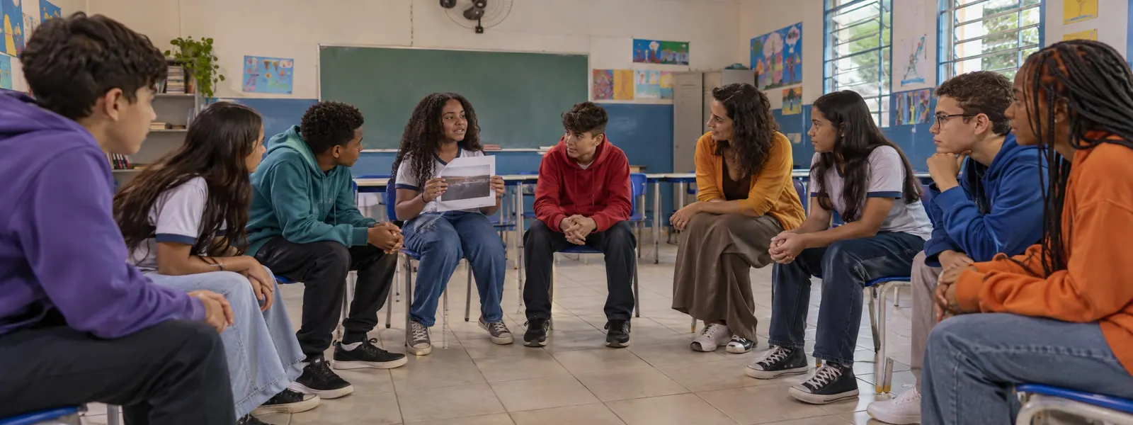 Estudantes sentados em roda na sala de aula, conversando com a professora